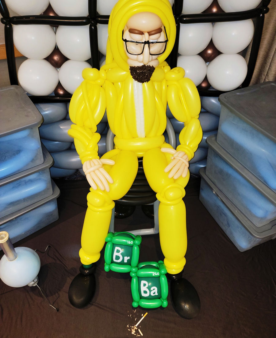 Life-size balloon sculpture of the Breaking Bad chemist in a yellow hazmat suit, seated beside balloon chemical-element blocks.