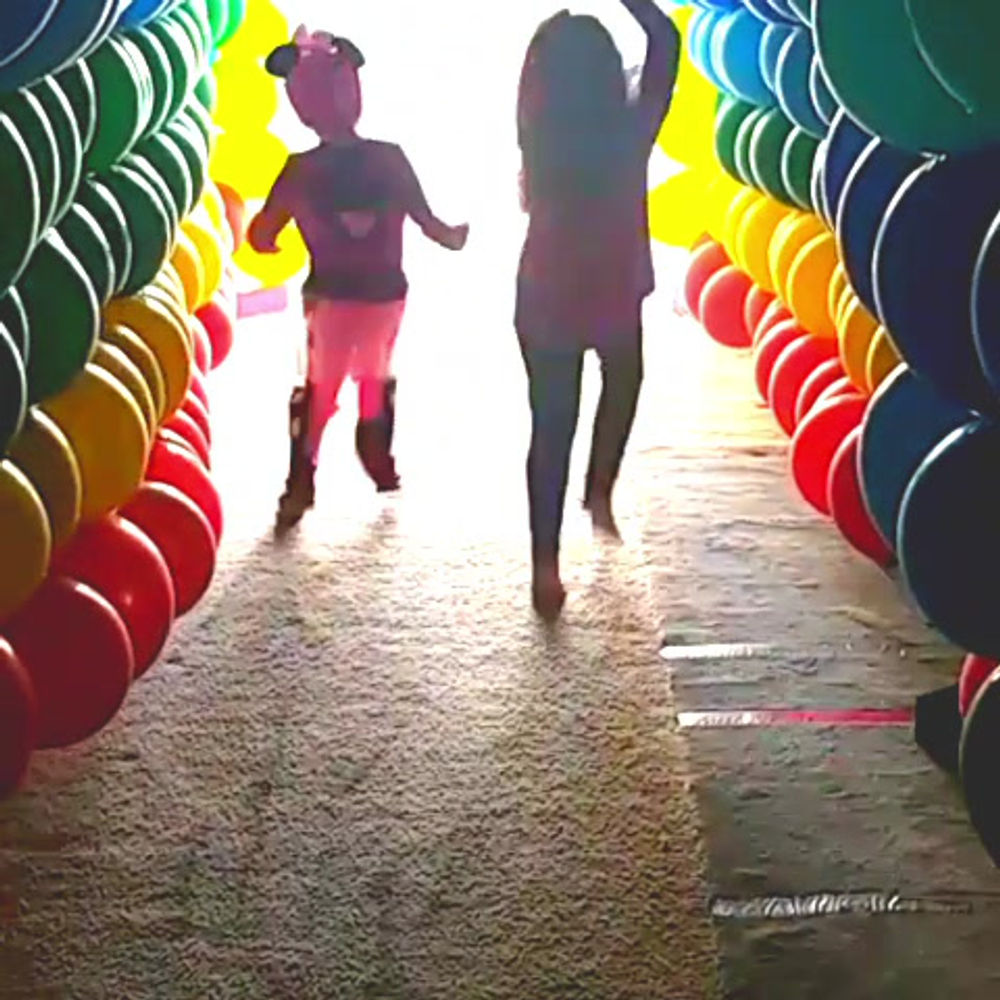 Children running through a long rainbow balloon tunnel toward daylight.