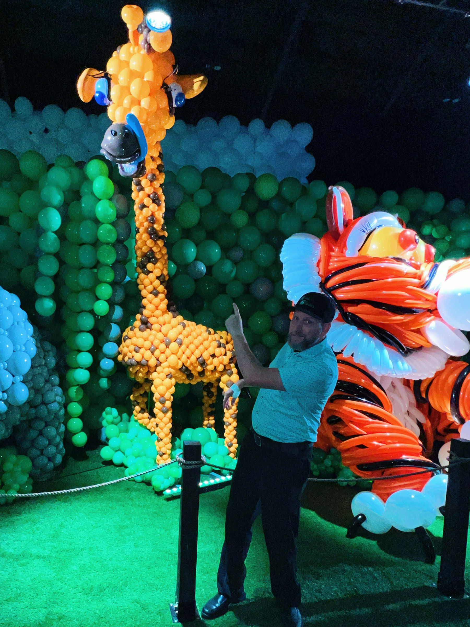 A walk-through balloon environment: a two-storey balloon giraffe and a giant balloon tiger built against a wall of green and blue balloons, with an artist standing beside them for scale.